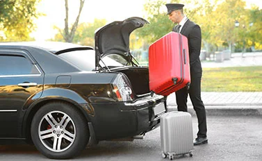 A chauffeur in a black suit and hat loading a red suitcase into the trunk of a black luxury car with a silver suitcase beside him on the pavement.